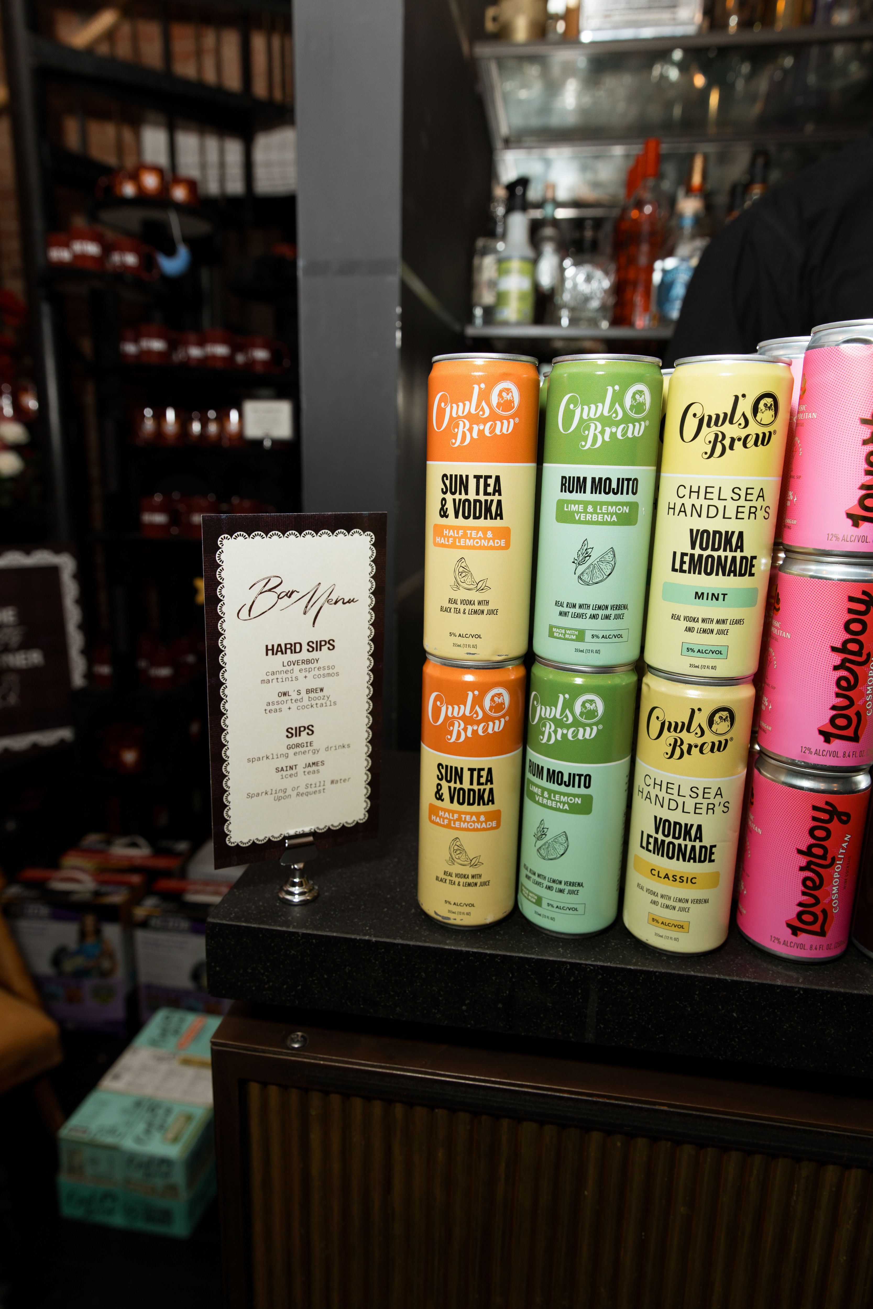Owl’s Brew canned cocktails and Loverboy drinks displayed beside the bar menu at ISR’s Social Impact House anniversary event.