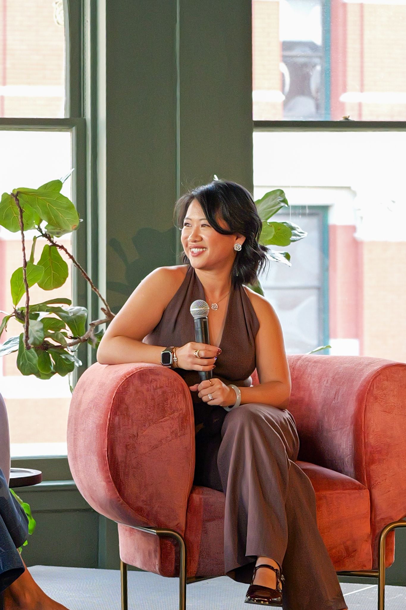Woman speaking on a panel while seated in a pink chair, holding a microphone and smiling.