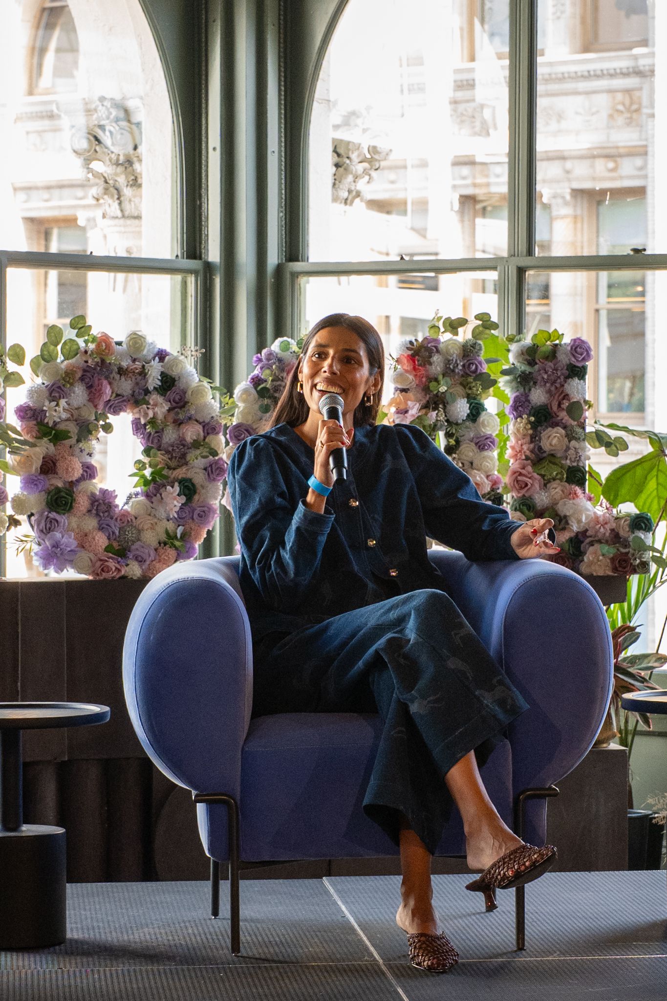 Woman speaking onstage at SoGal Summit, seated in a blue chair with a microphone and floral backdrop.