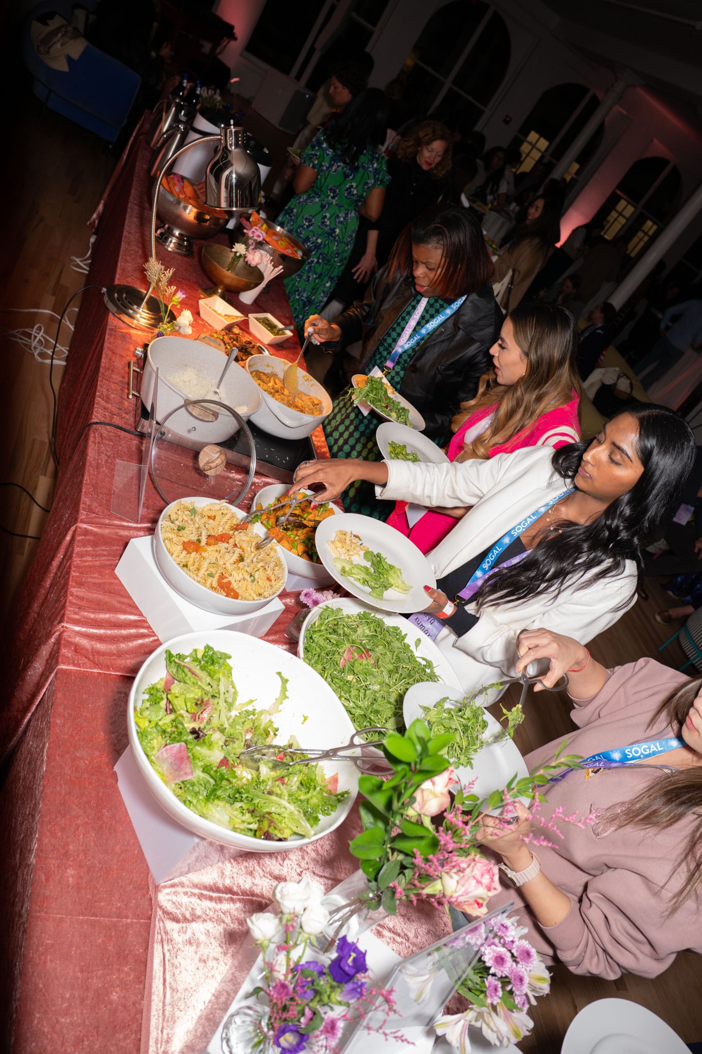 SoGal Summit guests serving dinner from a buffet with salads, warm dishes, floral arrangements, and SoGal lanyards.