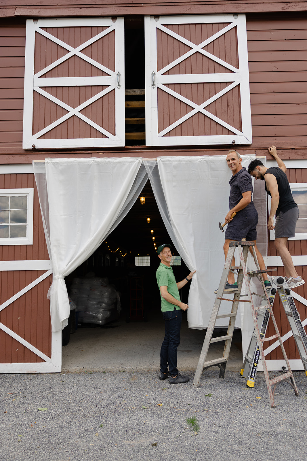 Three people hang white drapery across the entrance of a red barn at La Luna Farm while standing on ladders, preparing the space for an outdoor wedding celebration.